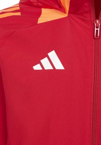 Veste rouge avec des accents orange, fabriquée en tissu lisse. Elle présente une fermeture éclair à l'avant et un logo blanc avec trois bandes sur la poitrine.