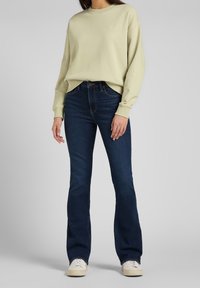 Sudadera de cuello redondo verde claro con puños acanalados, combinada con vaqueros acampanados azul oscuro. Zapatillas blancas informales completan el conjunto.