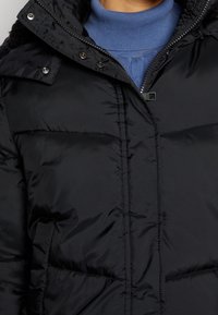 Chaqueta negra acolchada con diseño acolchado, cierre de cremallera, botones a presión y bolsillos laterales. La tela tiene una textura brillante y suave.