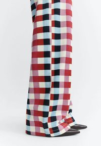Personne portant un pantalon à jambes larges avec un motif à carreaux rouge, bleu, noir et blanc, associé à des chaussures noires à bout pointu sur fond blanc.