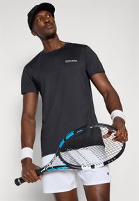 Juodas trumpomis rankovėmis sportinis marškinėlis su apvalia iškirpte, kuriame yra baltas logotipas ant krūtinės. Modelis laiko teniso raketę ir nešioja riešo juostas.