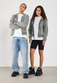 Due modelli stanno fianco a fianco. Uno indossa una felpa grigia con zip sopra una maglietta bianca, abbinata a jeans larghi azzurri e scarpe da ginnastica nere. L'altro indossa una felpa grigia abbinata, maglietta bianca, pantaloncini da bicicletta neri e scarpe da ginnastica nere.