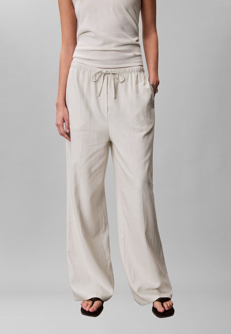 Άτομο που φοράει άνετο μπεζ παντελόνι με κορδόνι, άσπρο μπλουζάκι χωρίς μανίκια, με το ένα χέρι στην τσέπη και μαύρα πέδιλα ανοιχτά δάχτυλα σε γκρι φόντο.