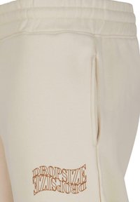 Beige joggebukser med snøring i livet, med en sidelomme og trykt "DROPSIZE" logo i brunt nær kanten. Myk tekstur.