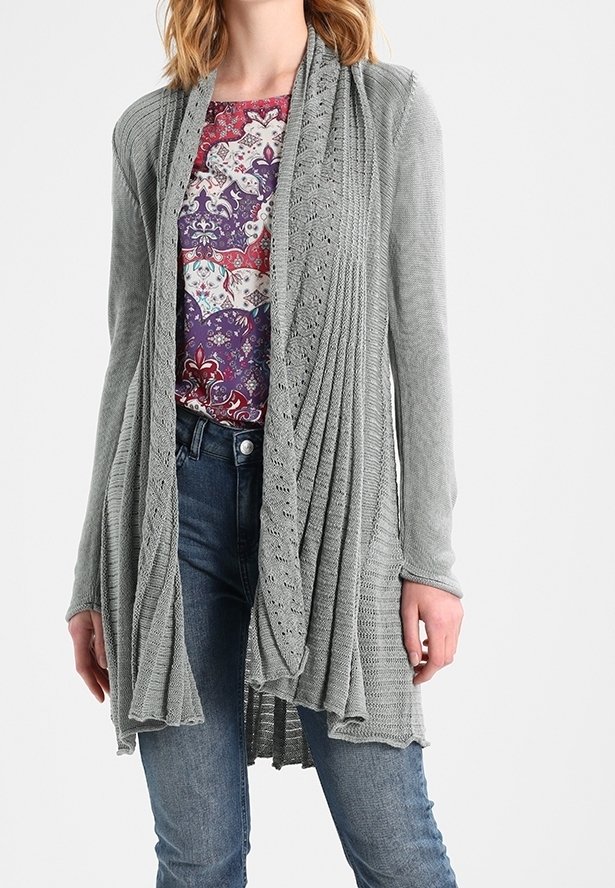 Femme portant un long cardigan gris en maille à ouverture sur le devant, sur un chemisier à motifs violets et rouges, avec un jean bleu, debout devant un fond blanc.