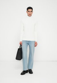 Pull à col roulé en tricot blanc, jeans droite bleu clair, chaussures en cuir noires et sac fourre-tout noir sur fond uni.