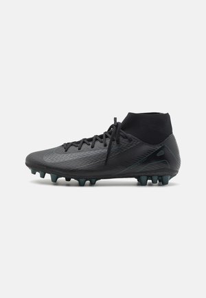 Chaussure de football montante noire avec lacets et crampons sur la semelle sur un fond gris clair uni.