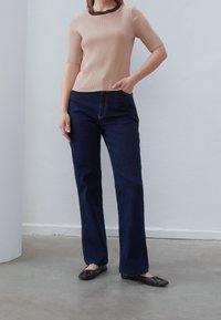 Beige žebrovaný top s krátkým rukávem a hnědým okrajem, kombinovaný s tmavě modrými rozšířenými džíny a černými plochými botami. Hladké textury, ležérní design.