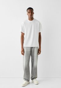 Witte t-shirt met korte mouwen gecombineerd met grijze wijde jeans. De jeans hebben een vervaagde textuur. Het model draagt witte sneakers.