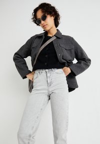 Casaco cinza em mistura de lã com dois bolsos no peito, suéter preto de botão, e jeans cinza claro de cintura alta. Óculos de sol redondos pretos completam o visual.