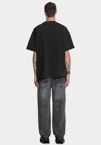 Czarna, oversize koszulka z okrągłym dekoltem, krótkim rękawem i gładką teksturą, w zestawieniu z szarymi, luźnymi dżinsami oraz czarnymi sneakersami z czerwonymi akcentami.
