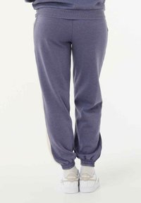 Persoon in paarse trainingsbroek en witte Reebok sneakers, staande met de voeten dicht bij elkaar op een lichte achtergrond.