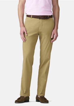 Mann trägt beigefarbene gerade geschnittene Hose, braunen geflochtenen Gürtel, braune Tassel-Loafer und ein hellrosa Hemd, das in die Hose gesteckt ist, steht vor weißem Hintergrund.