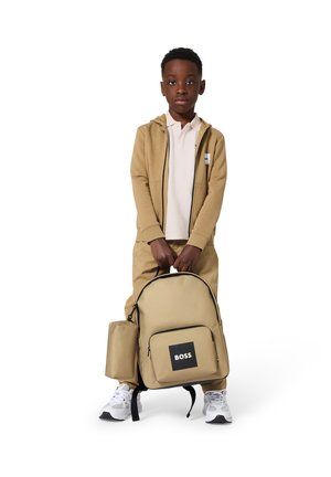 Jongen draagt een beige hoodie en broek, houdt een bijpassende beige rugzak vast met zwarte "BOSS"-logo, staand tegen een witte achtergrond.