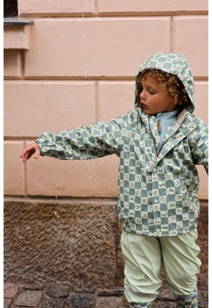 Zelená károvaná nepremokavá bunda s motívmi zvierat, doplnená o zodpovedajúce zelené nohavice a čižmy. Materiál je pravdepodobne nepremokavý; bunda má kapucňu.