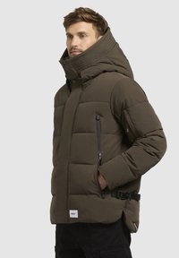 Puhasti kaput u tamno maslinastoj zelenoj boji, s visokim ovratnikom, zatvorenim džepovima i detaljem s kopčom sa strane. Kvadratna tekstura dodaje toplotnu izolaciju.