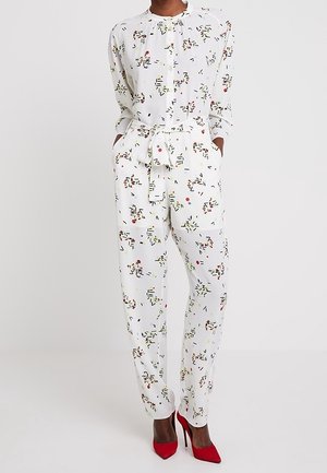 Žena v bílém dlouhém overal s vícebarevným abstraktním potiskem a stylovým vázáním v pase, doplněném jasně červenými vysokými podpatky.