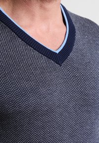 Mörk marinblå V-ringad tröja med ett fiskbensmönster, ribbad kant i marinblått och ljusblå accenter runt kragen. Mjuk struktur.