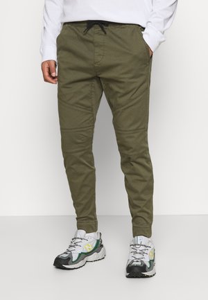 Mand iført olivengrønne joggerbukser med sort snor, hvide sneakers med grønne og gule detaljer, stående mod en hvid baggrund.