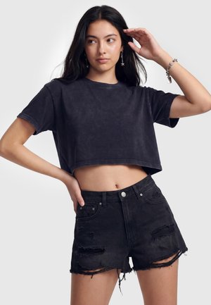 Kobieta ubrana w czarny krótki t-shirt i zniszczone czarne denimowe szorty stoi z jedną ręką na biodrze, a drugą podniesioną blisko głowy.