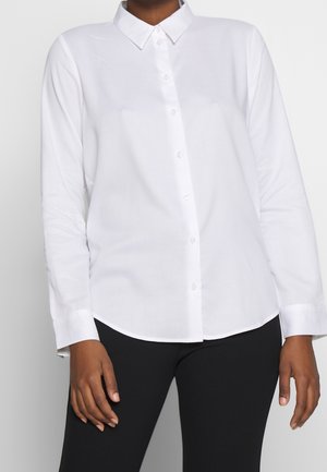 Osoba ubrana w długą białą koszulę na guziki z kołnierzykiem i czarne spodnie, stojąca na jednolitym jasnym tle.