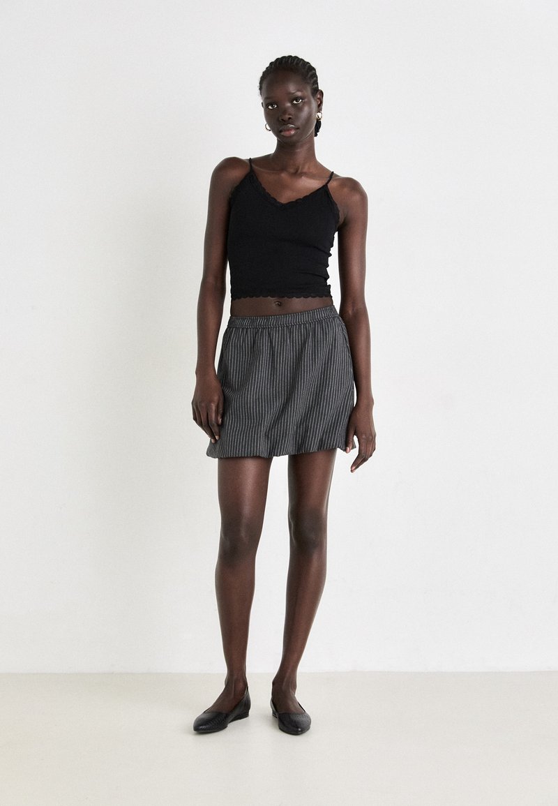 Schwarzes kurzes Tanktop mit gewellten Säumen, kombiniert mit einem grauen, gestreiften Minirock. Das Model trägt schwarze Ballerinas und steht vor einem neutralen Hintergrund.