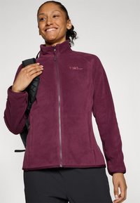 Giacca in pile color bordeaux con colletto alto, zip frontale completa e logo ricamato. Presenta una texture liscia e un design aderente, abbinata a uno zaino.