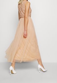 Yksipituinen mekko, jossa on paljetein koristeltu nude-vyötärö ja kerroksellinen läpikuultava tüllihame vaaleanbeigessä, yhdistetty valkoisiin korkokenkiin.