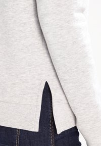 Sudadera gris clara con puños y dobladillo acanalados. Presenta aberturas laterales en la parte inferior y una textura suave. Combina con denim oscuro.