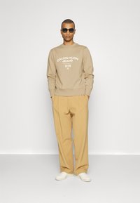 Felpa beige con testo bianco "CALVIN KLEIN JEANS 1978" abbinata a pantaloni beige larghi e sneakers bianche, indossata con occhiali da sole scuri.