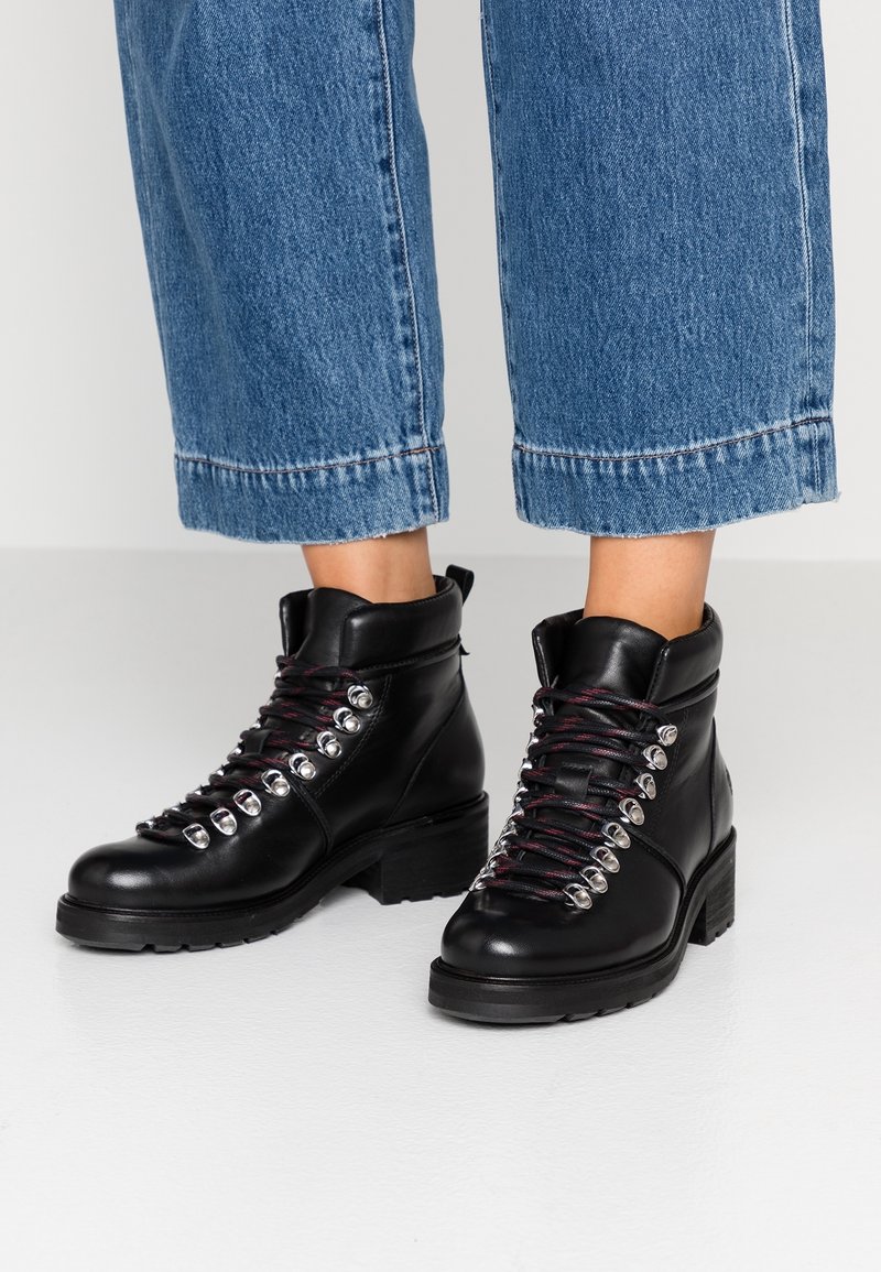 Persoon in zwarte leren enkellaarsjes met metalen veterogen en roodaccenten in de veters, gecombineerd met blauwe cropped denim jeans.