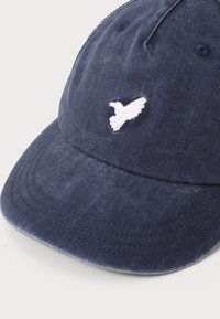Casquette bleu marine en tissu denim, avec un logo d'oiseau blanc brodé sur le devant, un visière courbée et des détails de couture visibles.