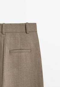 Pantalon beige sur mesure avec un tissu texturé, une ceinture lisse et une poche arrière unique. Comprend des passants pour ceinture et un design épuré et minimaliste.