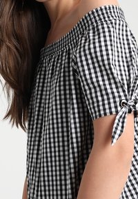 Black and white gingham off-the-shoulder blouse with short sleeves, pleated design, and ties on sleeve ends for adjustable fit.