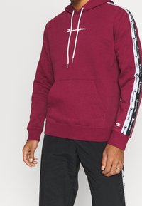 Uomo che indossa un hoodie maroon di Champion con cordini bianchi e nastro con logo sulle maniche, abbinato a pantaloni neri con strisce laterali.