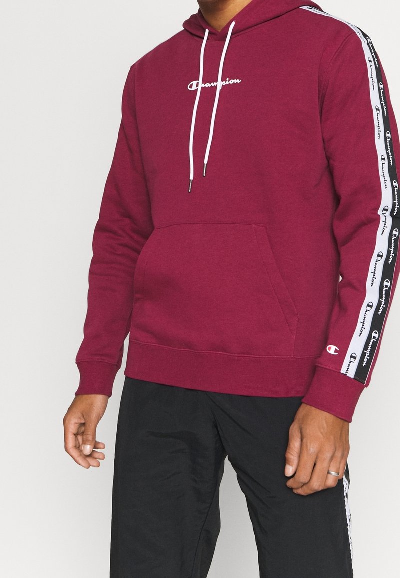 Uomo che indossa un hoodie maroon di Champion con cordini bianchi e nastro con logo sulle maniche, abbinato a pantaloni neri con strisce laterali.