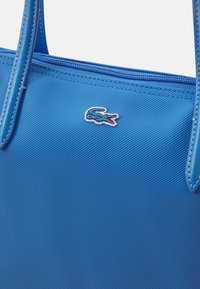 Sac fourre-tout en textile bleu avec fermeture éclair, anses en cuir lisse et petit logo crocodile blanc et bleu.