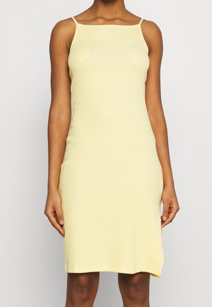 Femme portant une robe ajustée sans manches, longueur genou, jaune pâle, à fines bretelles sur un fond clair uni.