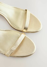 Gouden sandalen met banden hebben een gladde metallic afwerking, een platte zool en een minimalistisch ontwerp, met twee dunne bandjes bovenop.
