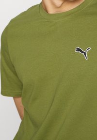 Personne portant un t-shirt vert olive avec un petit logo Puma noir et blanc sur le côté gauche de la poitrine.