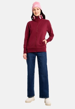 Frau, die eine pinke Mütze, eine burgunderrote Jacke mit Reißverschluss und Logo am Kragen, blaue Jeans und weiße Plateauschuhe trägt, steht mit einer Hand in der Tasche.