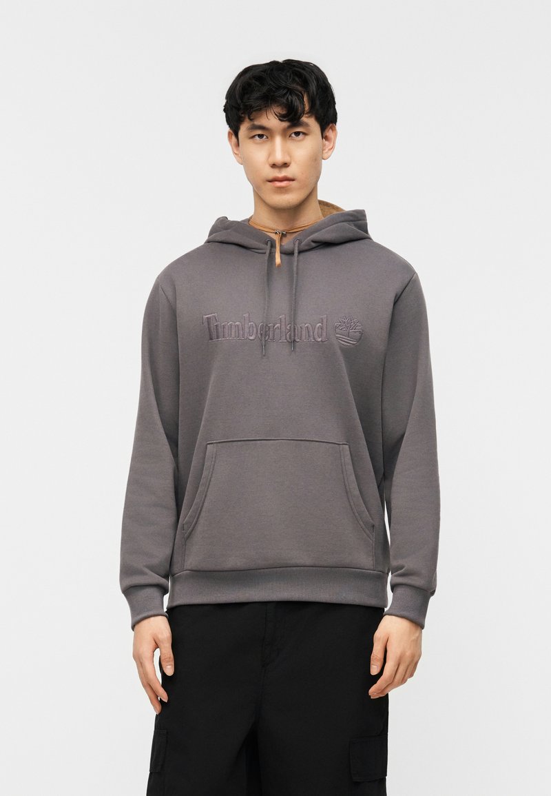 Νέος άνδρας που φοράει γκρι φούτερ Timberland με τσέπη καγκουρό και μαύρο παντελόνι cargo, στέκεται μπροστά σε απλό λευκό φόντο.