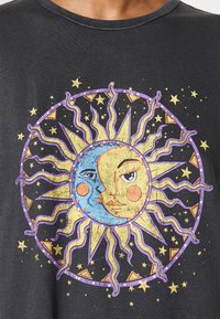 Black t-shirt featuring a central graphic of a sun and moon face, with yellow, blue, and purple details surrounded by stars and wavy rays.