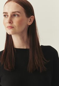 Personne aux cheveux bruns portant un pull noir avec un col côtelé. L'arrière-plan est neutre et de couleur claire.