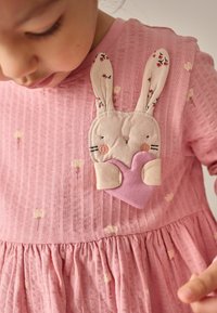 Růžové šaty s texturovanou tkaninou, s aplikací králíčka s dlouhýma ušima a srdíčkem. Jemný volánkový lem a malé květinové vzory.