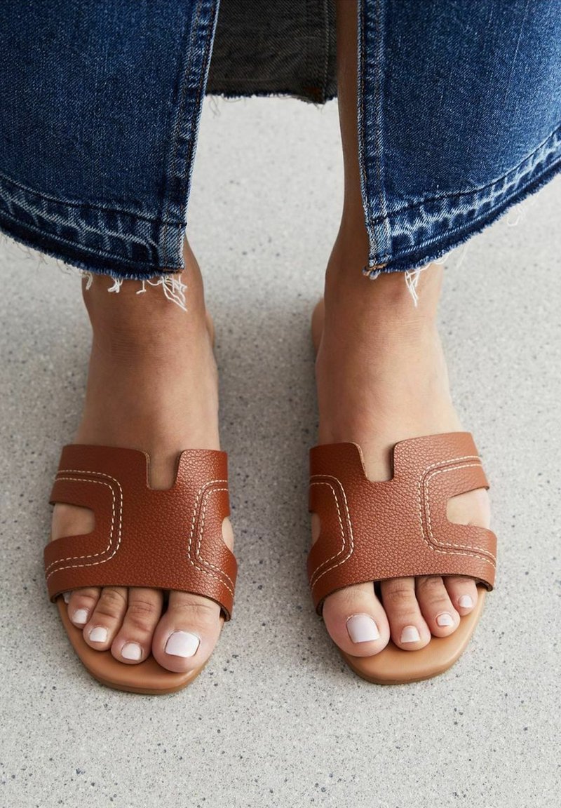 Sandali slide in pelle marrone con design a taglio, texture morbida e cuciture a contrasto. Suola piatta che mostra le dita dei piedi con uno smalto neutro, su una superficie di cemento.