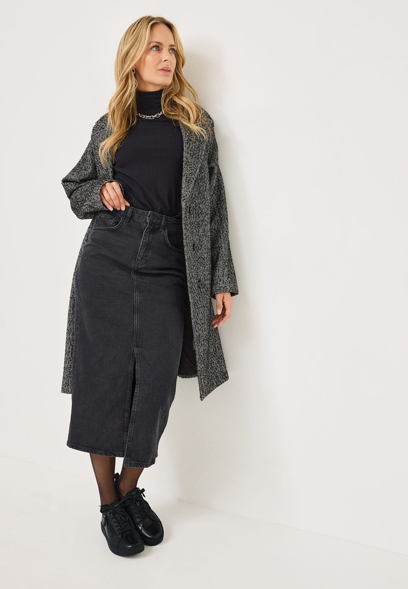 Un long cardigan gris texturé porté par-dessus un col roulé noir à côtes, associé à une jupe midi en denim noire avec une fente avant et des baskets noires.
