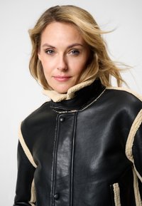 Zwarte leren jas met beige schapenvacht accenten, hoge kraag en drukknoopsluiting. Gevoel met een gladde textuur en contrasterende details.