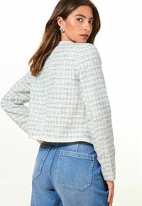 Crochetová bunda v světle modré a krémové barvě s texturovaným kostkovaným vzorem. Má dlouhé rukávy a zkrácenou délku, ve spojení s džíny.