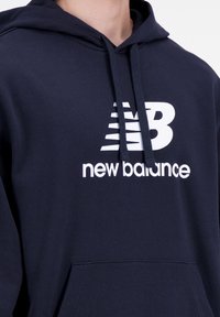 Felpa con cappuccio blu navy realizzata in tessuto morbido, con il logo bianco "New Balance" e cordini. Include una tasca frontale e polsini a costine.